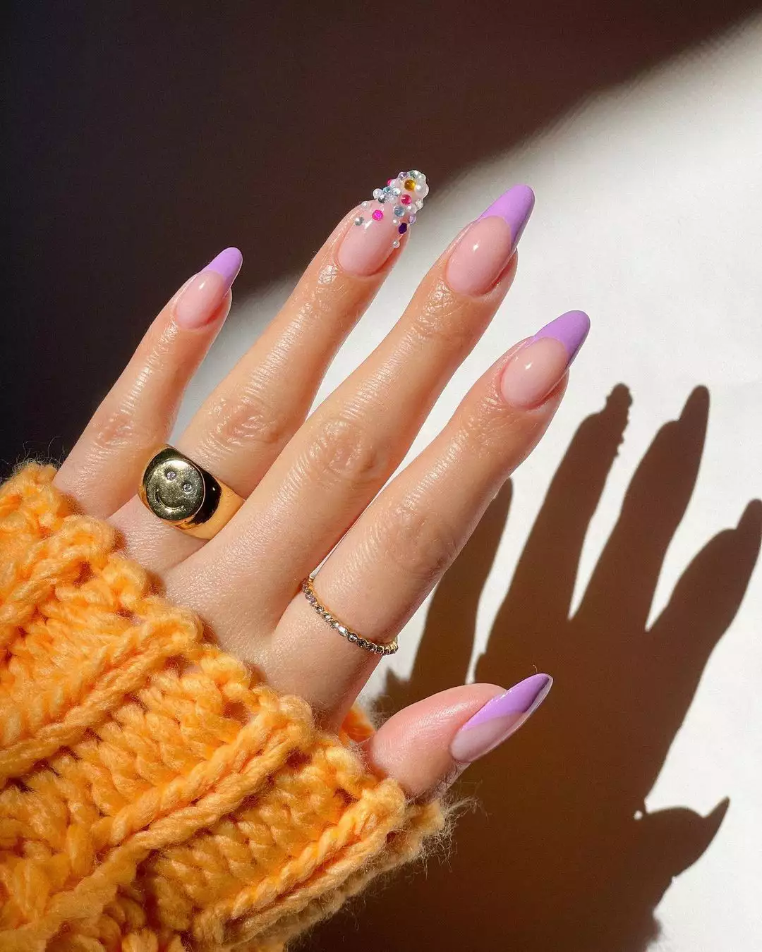 A hand adorned with rings and nail art with light purple nails and a single nail detailed with gems