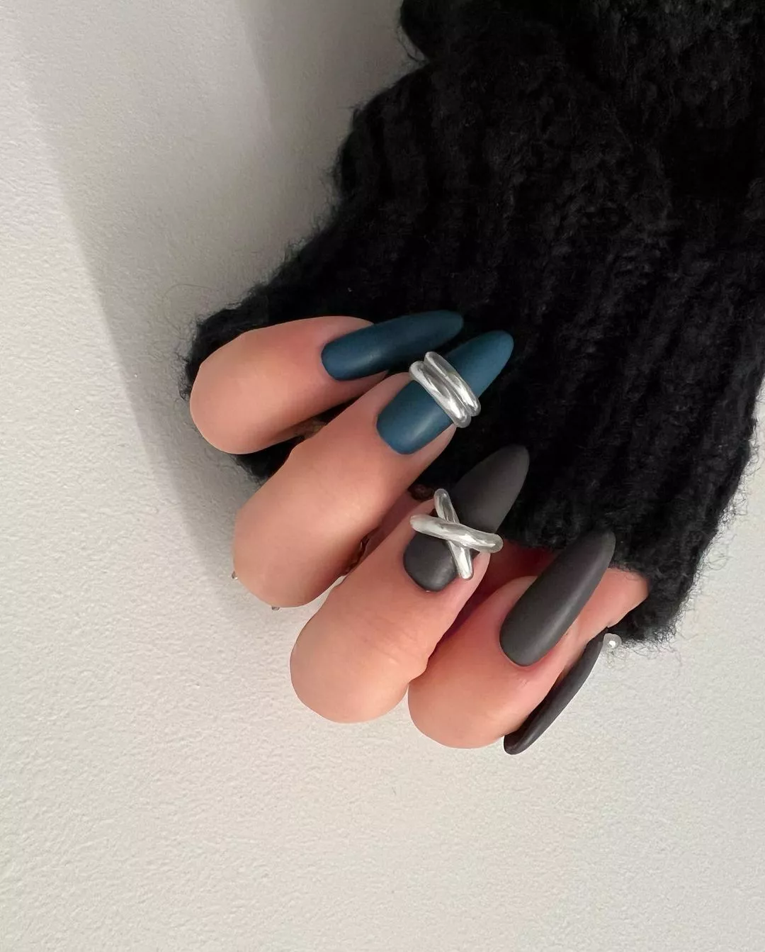 A hand with long matte nails decorated with silver rings wearing a black sweater