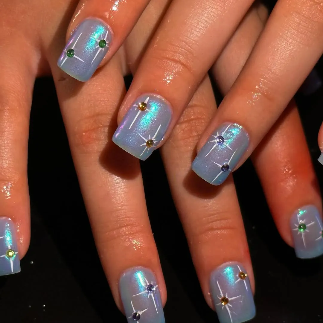 Short, square-shaped nails painted in a glittery, icy blue with colorful gems on each nail