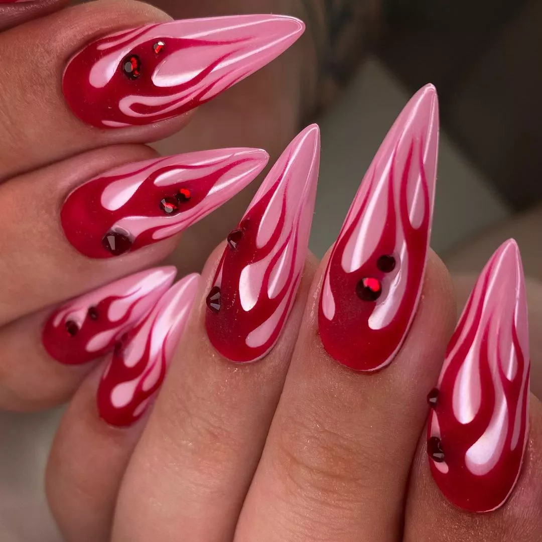 Person with stiletto-shaped nails that are metallic pink and have matte red flames and gems