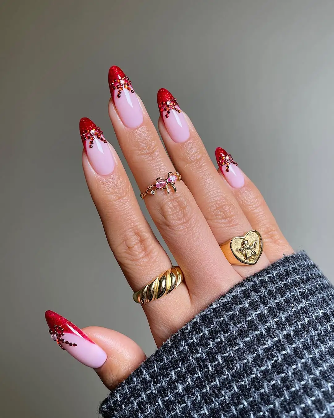 A hand displaying red and pink painted nails with nail art adorned with gold rings wearing a textured sleeve