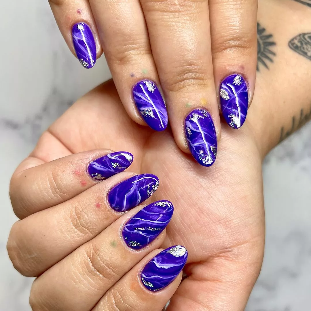 Person shows off their oval-shaped nails painted in a marbled royal purple with silver foil