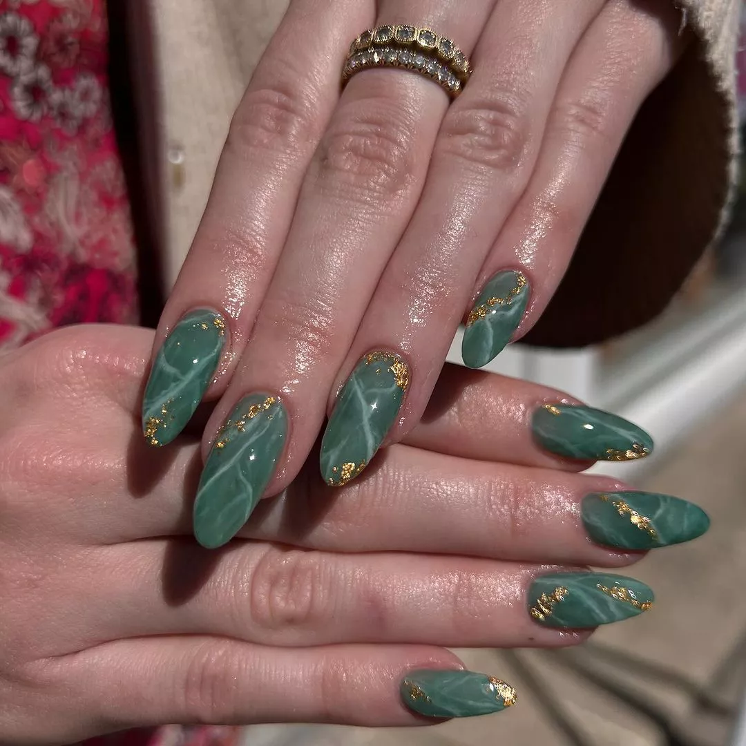 Person with long, oval-shaped nails painted in marble jade green and adorned with gold foil