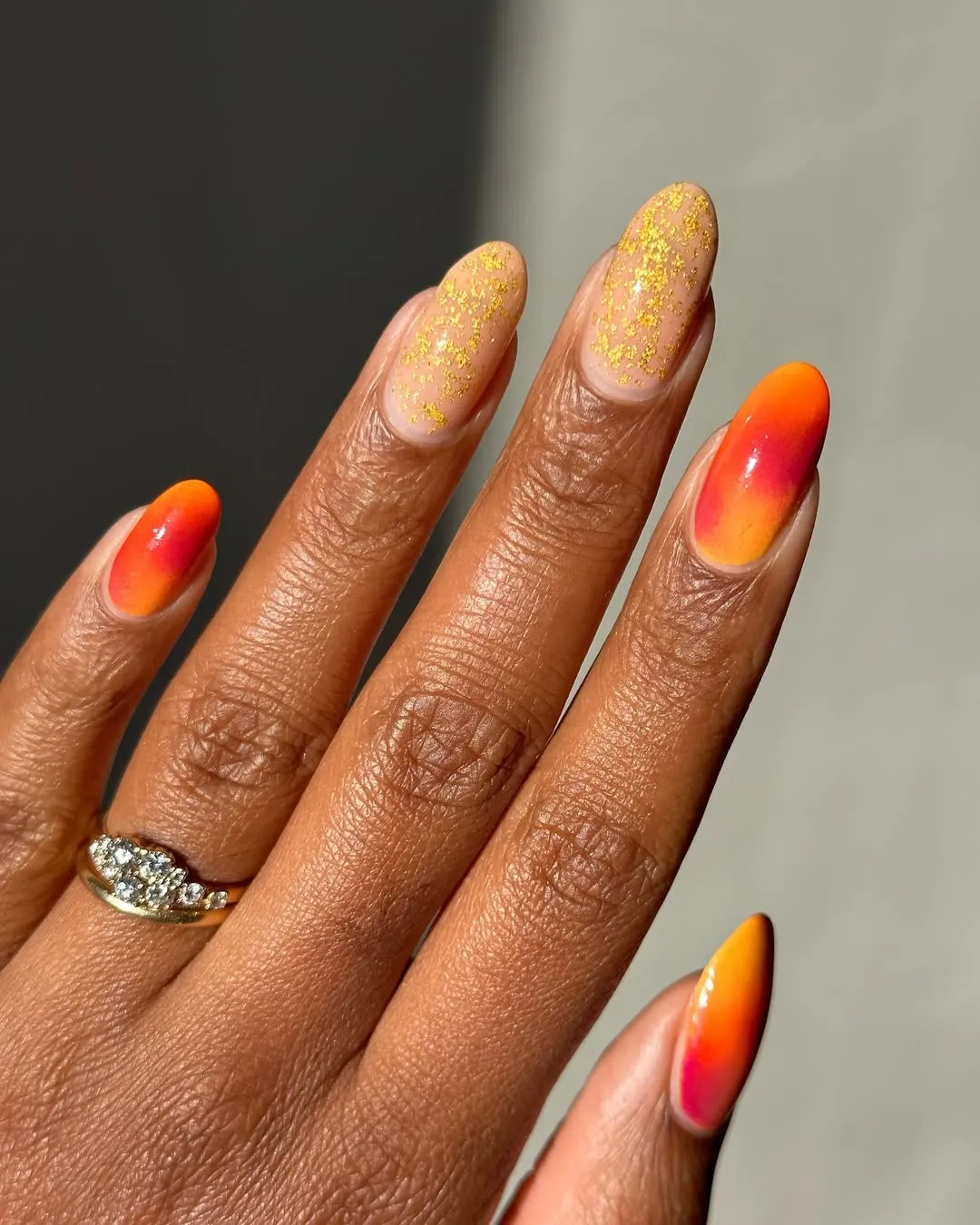 Person with oval-shaped nails that are red and orange hues with golden foil accents on two nails