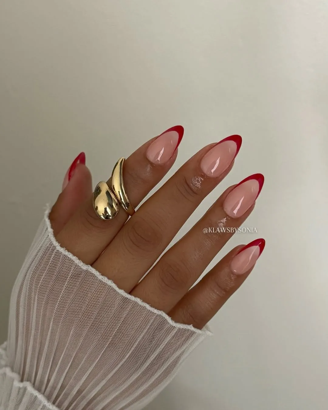 Hand with manicured nails featuring red tips wearing a gold ring and partially covered by a white sleeve