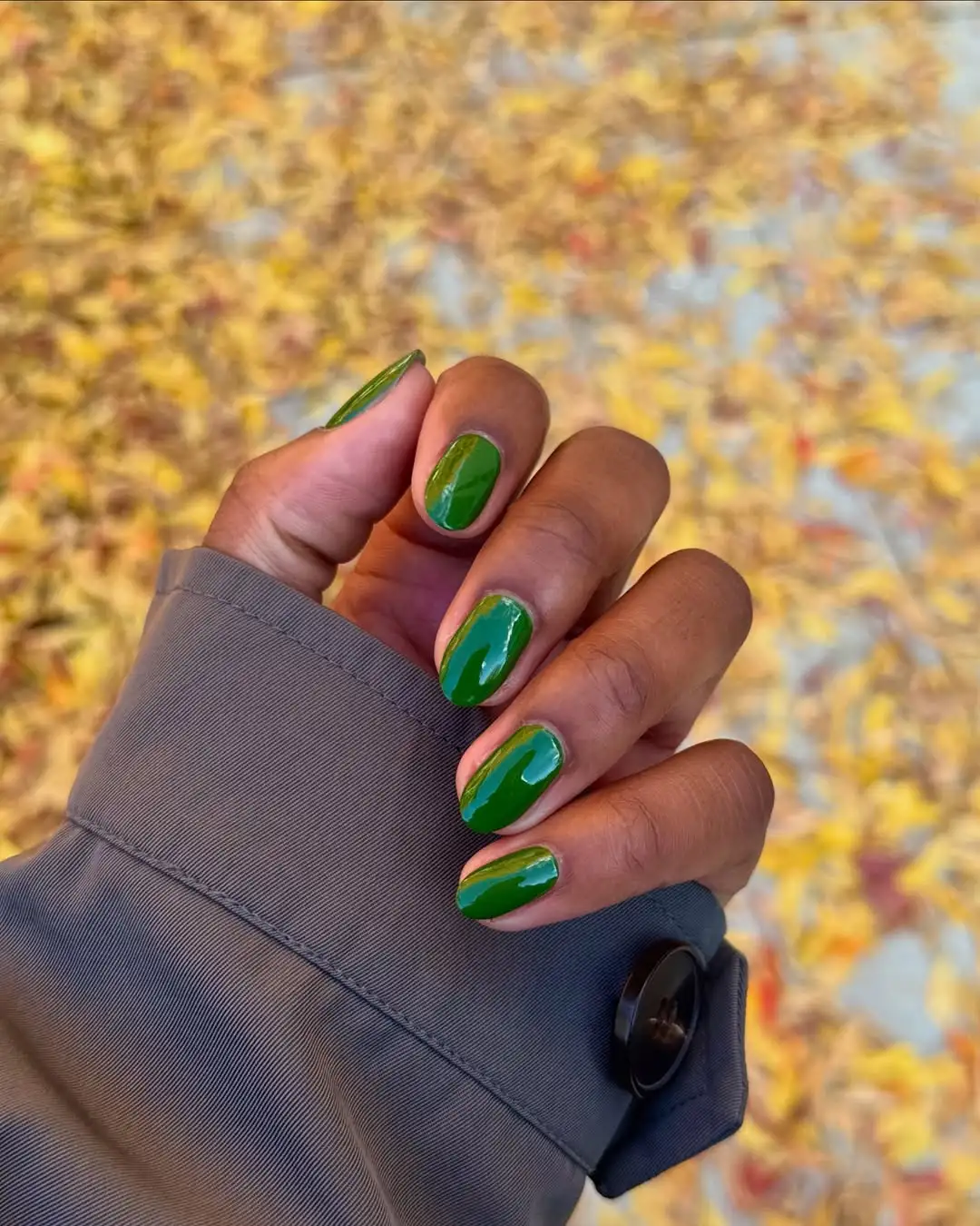Person holds their short, oval-shaped, green nails against a pile of yellowing leaves on the ground