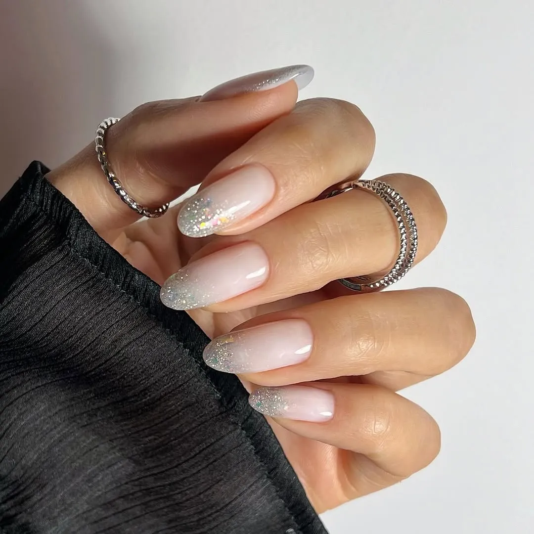 A hand wearing silver rings nails manicured with a gradient and glitter pattern