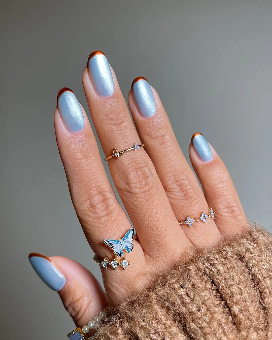 A hand with light metallic blue nail polish and redtipped nails adorned with rings including one with a butterfly design