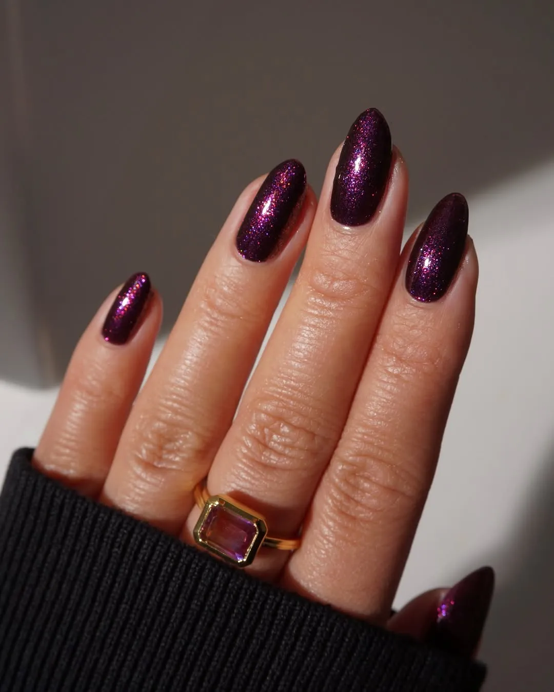 A closeup of a hand with long nails painted with a glittery finish wearing a ring with a rectangular purple stone