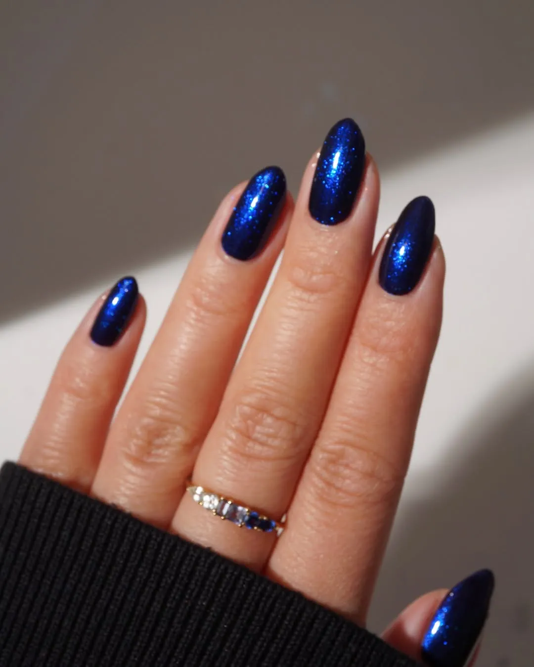 A hand with polished nails in a dark blue shade wearing a golden ring with small gemstones sleeve partially visible