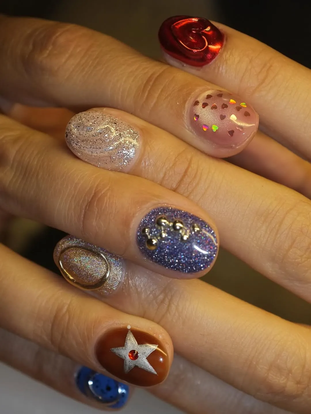 A closeup of hands with decorated fingernails displaying assorted nail art designs with various textures and elements