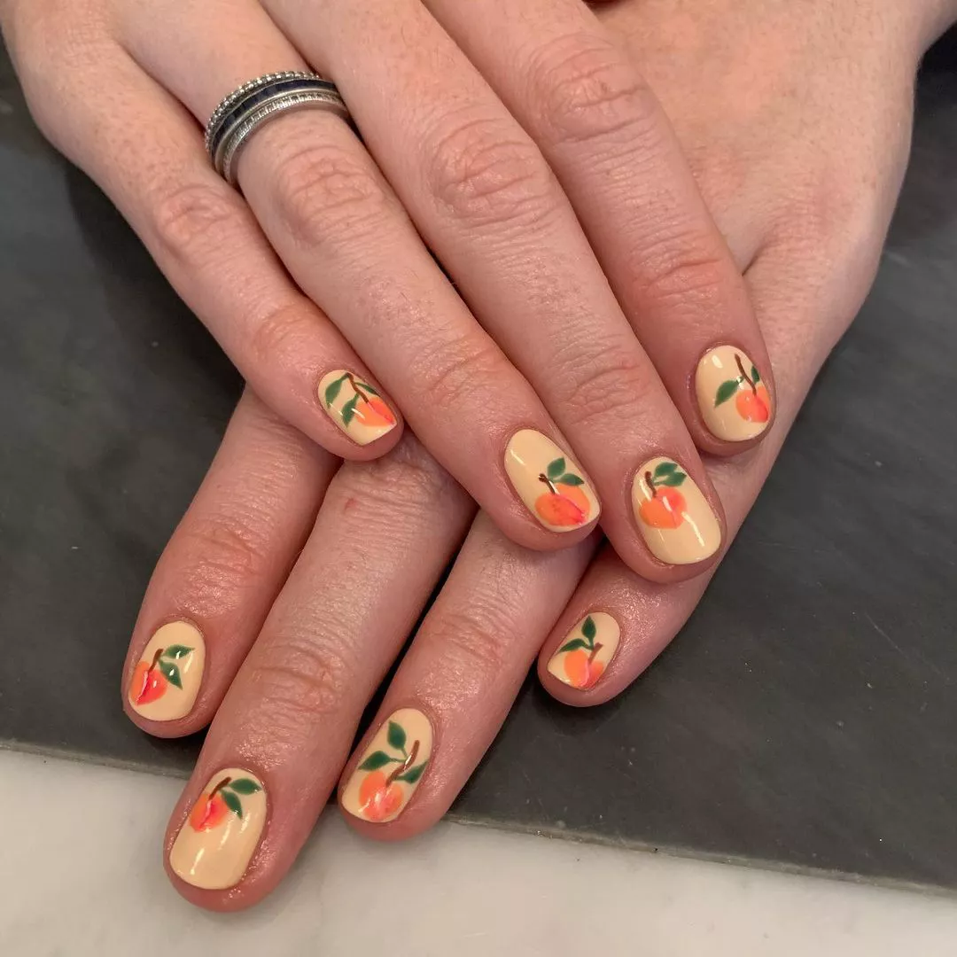 Person with oval-shaped nails painted in light peach with hand-painted peaches on each nail