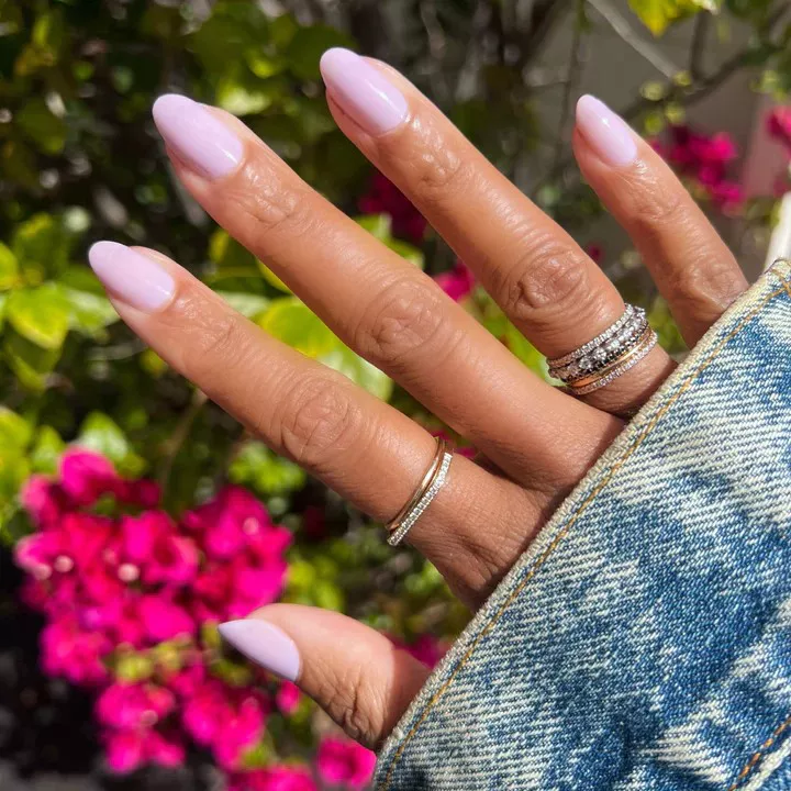 Person shows off their oval-shaped, sheer pink nails against a lush green and pink background