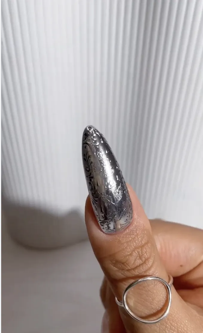 A hand wearing a silver ring showcasing a metallic patterned long fingernail