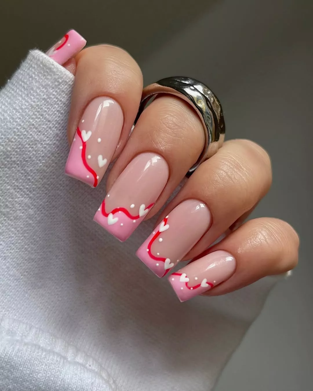 Person with square-shaped nails featuring pink French tips, red squiggly lines, and white hearts